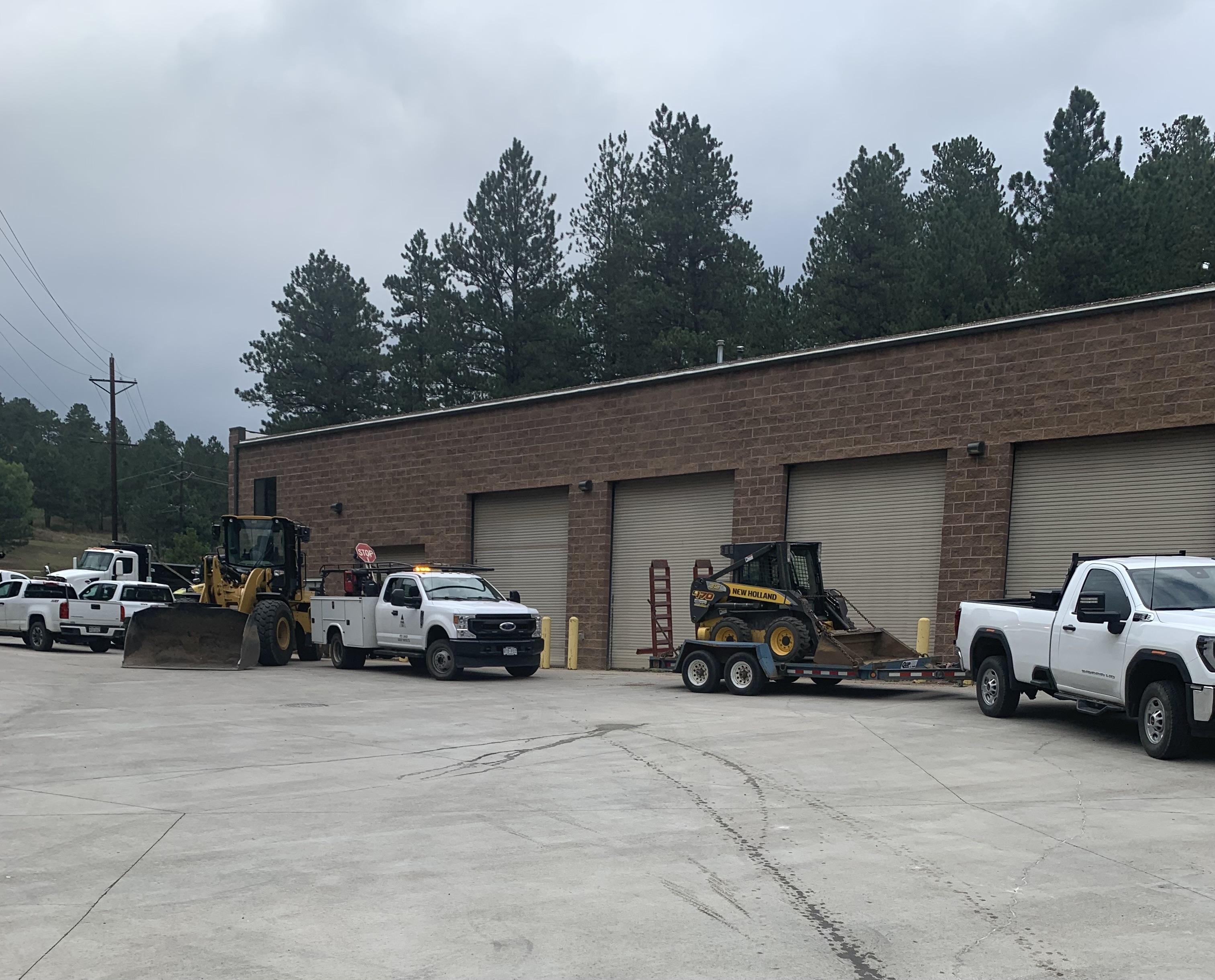 Picture of two white trucks, one with a skid steer loaded on a trailer. The vehicles are parked in front of the C&D building.